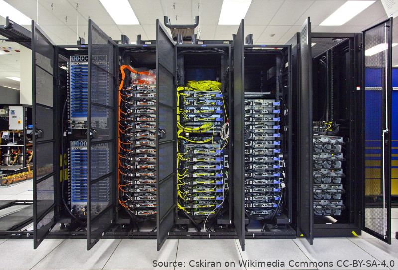 A photo of some high performance computer hardware racked in five cabinets in a server room. Each cabinet is about 2.2 metres high and 0.8m wide. The doors of the cabinets are open to show the systems inside. Orange and yellow cabling is prominent, connecting ports within the second and third racks.